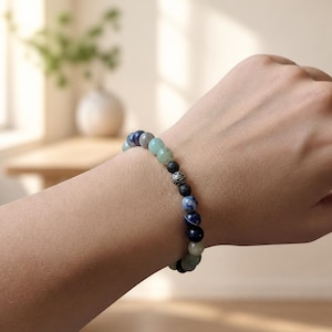 May include: A beaded bracelet featuring a mix of round polished stones in shades of blue, green, and grey, interspersed with textured black lava beads and a single silver-toned metal spacer bead with an intricate engraved pattern.