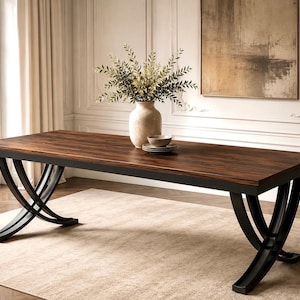 Pode incluir: Uma mesa de jantar retangular com tampo de madeira castanho-escura e base de metal preto com dois conjuntos de pernas curvas e cruzadas. Um vaso de cerâmica bege com folhagem e uma pequena pilha de tigelas estão sobre a mesa.