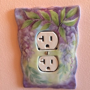 以下が含まれることがあります： 紫色のフジの花と緑の葉が描かれた、装飾的な磁器製のコンセントカバー。カバーには標準的なコンセントが2つあります。