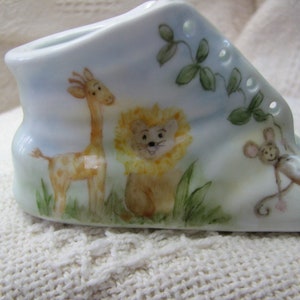 May include: A white ceramic baby shoe with a blue and white background. The shoe is decorated with a giraffe, a lion, and a mouse.