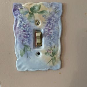 Op de afbeelding: Een porseleinen lichtschakelaar met een bloemenmotief. De afdekking is geschilderd met een lichtblauwe achtergrond en paarse bloemen. De schakelaar is gemarkeerd met "ON".