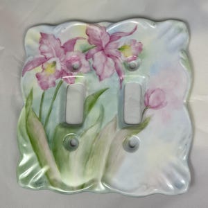 May include: A decorative double light switch cover with a hand-painted floral design. The cover has a scalloped edge and is painted with pink orchids, green leaves, and a soft pastel background.