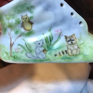 May include: A small, white ceramic baby shoe with hand-painted woodland creatures. An owl, squirrel, and raccoon are depicted in a grassy scene with trees and flowers. The shoe has eyelets for laces and is a charming keepsake.