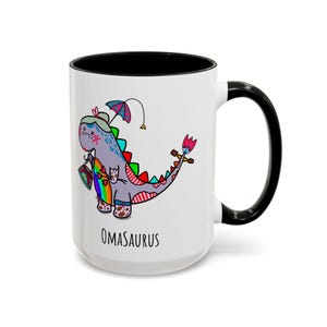 May include: A white ceramic mug with a black interior and handle, featuring a colorful illustration of a dinosaur character holding a handbag and an umbrella, with the text OMASAURUS printed in black lettering below the graphic design.