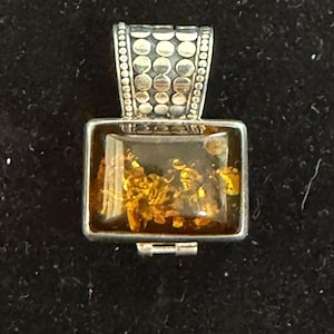 Peut inclure: Un pendentif rectangulaire composé d'une pierre d'ambre polie aux inclusions dorées, sertie dans un cadre en argent. La bélière est ornée d'un motif vertical de cercles en argent en relief et de bords perlés, contrastant avec la pierre lisse.