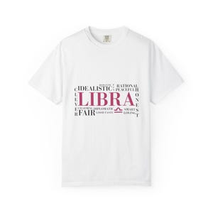 Può includere: T-shirt bianca con la parola "LIBRA" in rosa, circondata da parole come "leale" e "amorevole". È incluso anche il simbolo della Bilancia. Girocollo.