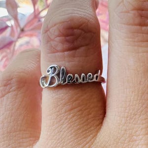 May include: A silver-toned ring featuring the word Blessed in a cursive script font. The ring is worn on a finger, showcasing the metallic finish and the stylized lettering that forms the band. The design is simple, with the text integrated into the ring.