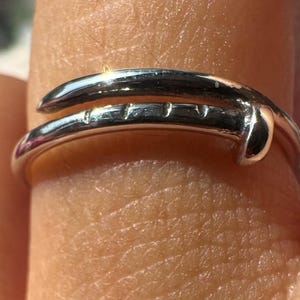 May include: A polished silver-tone ring shaped like a bent nail that wraps around the finger. The design features a rounded head at one end and a tapered point at the other, with small etched markings along the shank of the metal band.