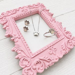 May include: A pink, ornate picture frame displays a silver paw print necklace, ring, and stud earrings. The jewelry is set against a white background. The frame has a detailed, carved design.