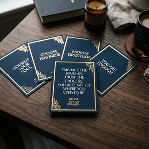 May include: A collection of navy blue affirmation cards with gold accents and inspirational quotes, such as "Nourish Your Soul" and "Choose Kindness." The cards are arranged on a dark wooden surface, alongside a cup of coffee and a candle.