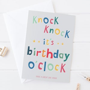 Puede incluir: Tarjeta de felicitación de cumpleaños con el texto "Knock Knock it's birthday o'clock" en letras coloridas. La tarjeta es de color gris claro e incluye el mensaje "Have a great day, Ryan!". Un sobre blanco está al lado.