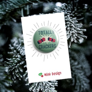 May include: A green button with the text "TOTALLY CRACKERS" and a cracker graphic, on a white card. The card has the "Wink Design" logo. The background features frosted evergreen branches, evoking a festive Christmas theme.