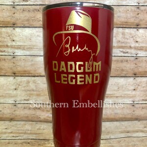 May include: A red, stainless steel tumbler with a gold inscription that reads "FSU, Bobby,  Dadgum Legend". The tumbler is on a wooden surface.