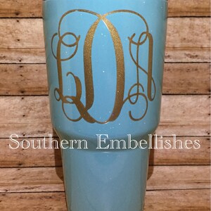 May include: A turquoise glitter tumbler with a gold monogram "LDM" on the side. The tumbler is sitting on a wooden surface.