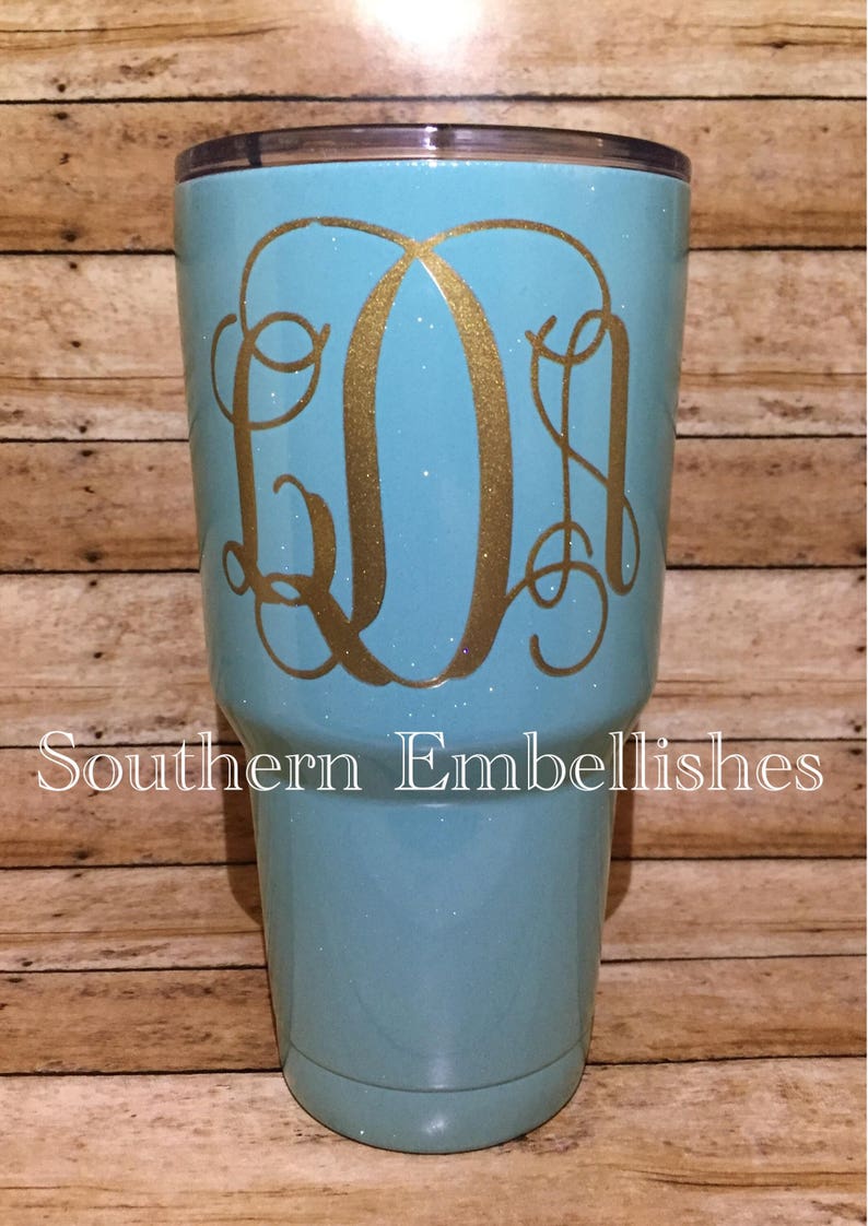 May include: A turquoise glitter tumbler with a gold monogram "LDM" on the side. The tumbler is sitting on a wooden surface.