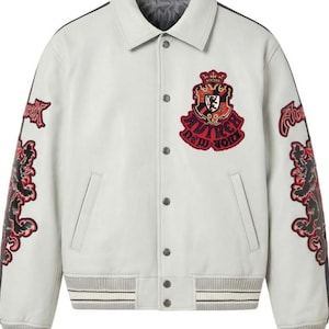 Puede incluir: Chaqueta universitaria gris claro con parches bordados en negro y rojo. La chaqueta tiene una parte delantera abotonada, un cuello y pu&ntilde;os y dobladillo acanalados con rayas blancas. Los parches incluyen un escudo y el texto "Avirex New York".