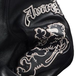 Puede incluir: Manga de chaqueta de cuero negro con parches bordados. Los parches presentan la palabra "Avirex" y un emblema de le&oacute;n, ambos con contornos blancos.