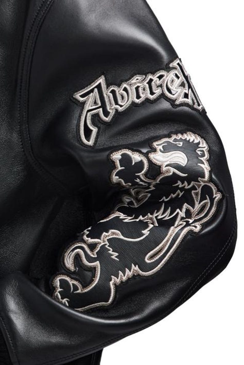Puede incluir: Manga de chaqueta de cuero negro con parches bordados. Los parches presentan la palabra "Avirex" y un emblema de le&oacute;n, ambos con contornos blancos.