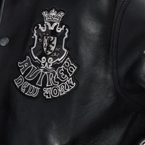 Puede incluir: Chaqueta de cuero negra con un parche decorativo con un escudo, una corona y las palabras "AVIREX NEW YORK" en blanco. La chaqueta tiene cierre de bot&oacute;n y cuello.
