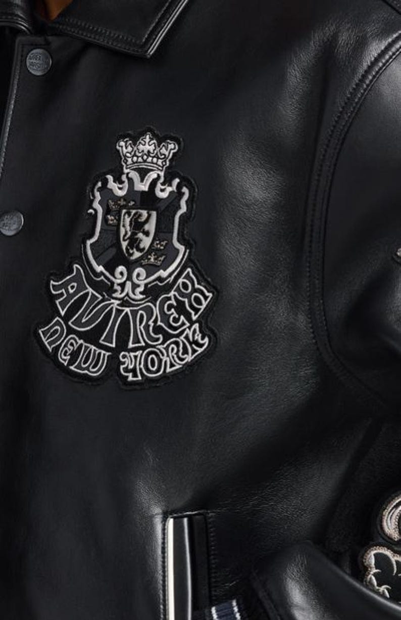 Puede incluir: Chaqueta de cuero negra con un parche decorativo con un escudo, una corona y las palabras "AVIREX NEW YORK" en blanco. La chaqueta tiene cierre de bot&oacute;n y cuello.