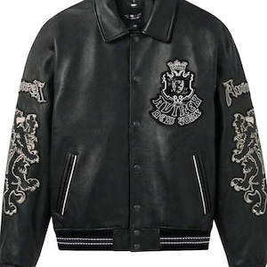 Puede incluir: Chaqueta de cuero negro con cuello cl&aacute;sico y cierres a presi&oacute;n. Cuenta con parches bordados en las mangas y el pecho, incluyendo un escudo y el texto "Aviirex New York". La chaqueta tiene ribetes blancos y un dobladillo a rayas.