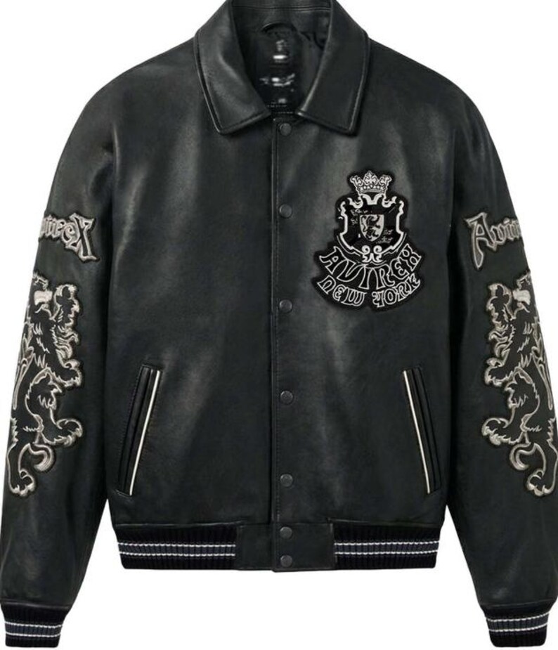 Puede incluir: Chaqueta de cuero negro con cuello cl&aacute;sico y cierres a presi&oacute;n. Cuenta con parches bordados en las mangas y el pecho, incluyendo un escudo y el texto "Aviirex New York". La chaqueta tiene ribetes blancos y un dobladillo a rayas.