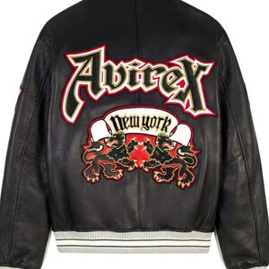 Puede incluir: Chaqueta de cuero negra con cuello y pu&ntilde;os blancos. La espalda presenta un gran dise&ntilde;o bordado con la palabra "Avirex" en rojo y dorado, y "New York" en una pancarta. Debajo, dos figuras de leones.
