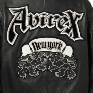 Puede incluir: Chaqueta de cuero negra con el nombre de la marca "Avirex" en letras blancas y negras en la espalda. Debajo, una pancarta blanca con el texto "New York" y dos emblemas de leones.