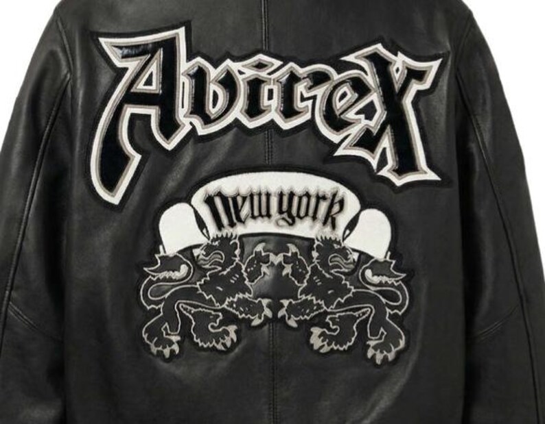 Puede incluir: Chaqueta de cuero negra con el nombre de la marca "Avirex" en letras blancas y negras en la espalda. Debajo, una pancarta blanca con el texto "New York" y dos emblemas de leones.