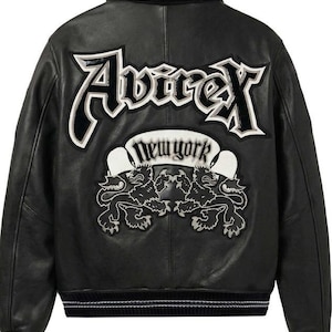 Puede incluir: Chaqueta de cuero negra con letras y parches bordados en blanco y negro. La parte trasera de la chaqueta presenta la palabra "Avirex" en letras grandes y estilizadas, y una pancarta que dice "New York" con dos emblemas de leones.