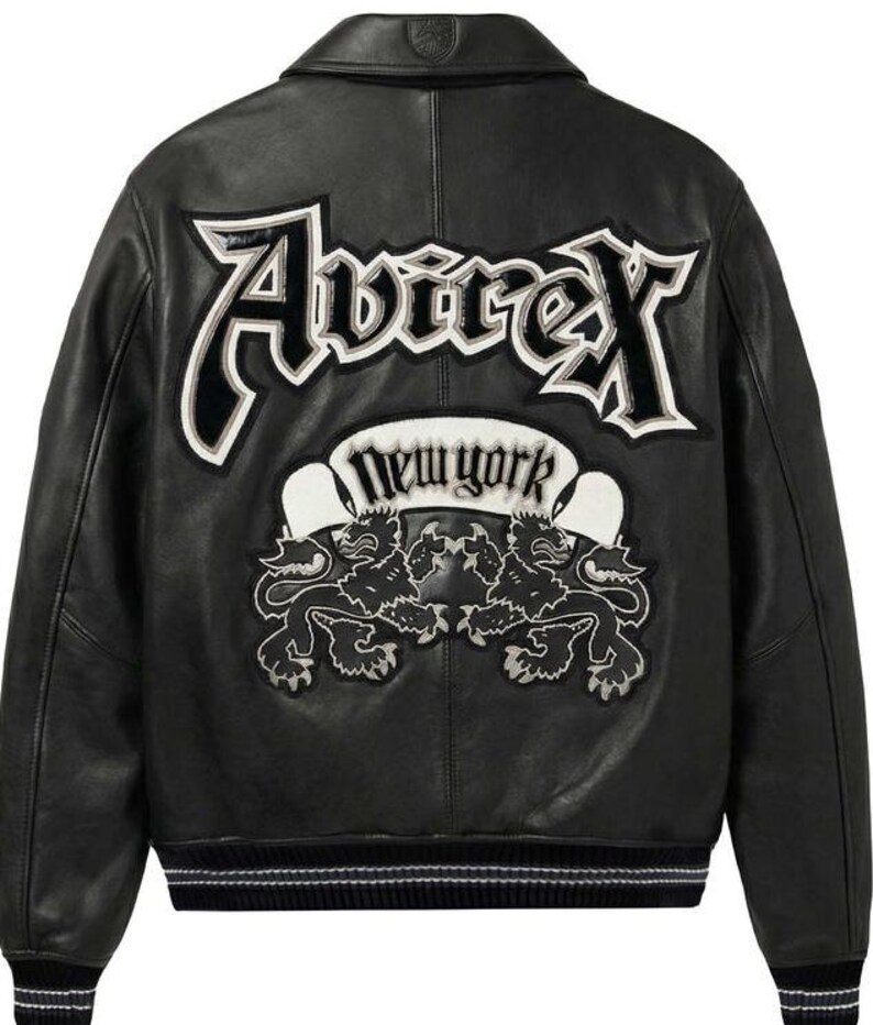 Puede incluir: Chaqueta de cuero negra con letras y parches bordados en blanco y negro. La parte trasera de la chaqueta presenta la palabra "Avirex" en letras grandes y estilizadas, y una pancarta que dice "New York" con dos emblemas de leones.