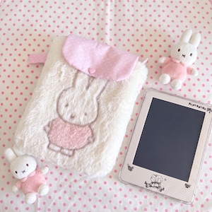 Puede incluir: Un estuche de piel sintética blanca con una solapa de lunares rosas y un botón en forma de corazón, con un bordado de Miffy. Dos pequeños peluches de Miffy con trajes rosas y una tableta digital con marco de lunares están colocados cerca.