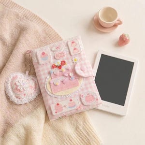 May include: A pink and white gingham fabric e-reader cover decorated with felt cake and dessert appliques, featuring a snap closure and a heart-shaped charm. Beside it sits a white e-reader, a small pink teacup on a saucer, and a strawberry figurine.