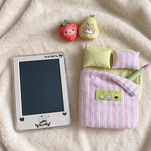 Könnte beinhalten: Ein digitales Tablet mit einem weiß gepunkteten Rahmen und kleinen schwarzen Illustrationen, ein Miniaturbett mit hellgrünem Untergrund, zwei Kissen, eine rosa-weiß gestreifte Decke sowie zwei kleine Plüschtiere in Form einer Erdbeere und einer Birne.
