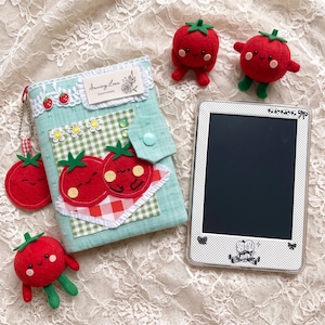 May include: A light blue fabric cover featuring a white lace trim, a label reading Sewing Love HANDMADE, and a green gingham patch with two red felt tomatoes. Includes a matching tomato keychain, three felt tomato figures, and an e-reader.