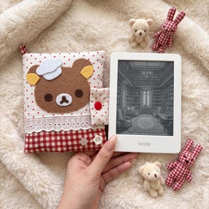 Pode incluir: Um leitor de e-book Kindle branco exibindo uma ilustração de biblioteca ao lado de uma capa de tecido com um aplique de urso marrom usando um chapéu de chef branco, tecido de bolinhas vermelhas, xadrez vermelho e dois botões.