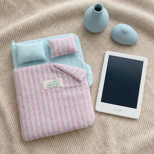 Pode incluir: Um conjunto de cama em miniatura com um colchão azul-claro, uma almofada azul-claro lisa e uma almofada às riscas cor-de-rosa e brancas. Inclui um edredão às riscas cor-de-rosa e brancas a condizer com uma etiqueta retangular com dois golfinhos e um Kindle branco.