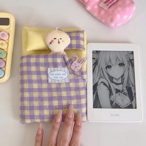Pode incluir: Uma capa para leitor eletrónico com xadrez amarelo e roxo, com uma pequena personagem de pelúcia num design de cama e uma etiqueta com a inscrição THE STARS ARE FOR YOU, ao lado de um leitor branco com uma personagem anime e uma bolsa rosa.