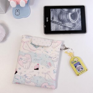 以下が含まれることがあります： 子羊、ケーキ、リボン、星のパステルカラーの模様が描かれた、フラップ付きの長方形の布製ポーチ。ポーチの側面には、パンダのイラストと「MY LUCK TODAY」という文字が入った黄色いプラスチック製のキーホルダーが取り付けられています。