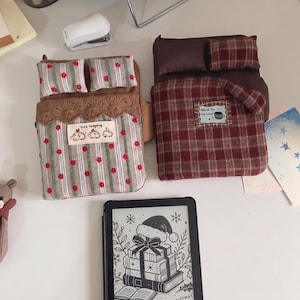 Pode incluir: Duas capas retangulares de tecido para e-readers projetadas como camas em miniatura com travesseiros e cobertores. Uma apresenta um padrão de listras cinzas com flores vermelhas e uma etiqueta "Cute hedgehog", a outra um padrão xadrez vermelho.