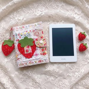 May include: A white e-reader with a black screen next to a fabric cover featuring a red felt strawberry appliqué, white lace trim, and a floral pattern. The cover includes a matching strawberry charm and a snap closure, with two small felt strawberries nearby.
