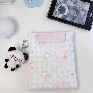 May include: A quilted fabric sleeve for an e-reader featuring a light blue and white gingham pattern with illustrations of cats, pink bows, and flowers. The sleeve has a pink polka-dot flap closure and a plush panda keychain with red glasses attached.