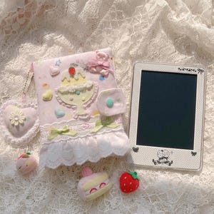 May include: A light pink fabric notebook cover decorated with a plush dessert applique, a bear charm, and lace trim, accompanied by a heart-shaped keychain, a small plush cake, a strawberry, and a digital tablet with a white polka dot frame.
