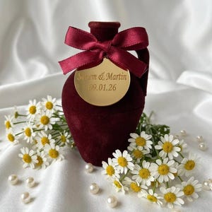 May include: A burgundy velvet vase with a gold tag and a burgundy ribbon. The tag reads "Sanem & Martin 09.01.26". White daisies and pearls surround the vase, creating a romantic and elegant display.