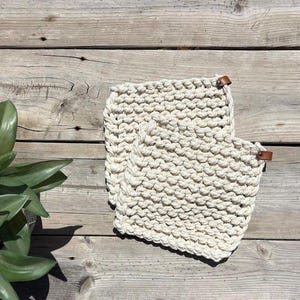 May include: Two square, off-white, hand-knitted potholders featuring a thick, textured garter stitch pattern. Each potholder is accented with a small, brown, rectangular leather tab attached to one corner with a metal rivet for hanging.