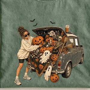 May include: A sage green t-shirt with a Comfort Colors label at the neck. The graphic features a person in a white sweatshirt and black shorts standing by a car boot filled with pumpkins, ghosts, a skeleton, and bats. The text reads Comfort Colors.