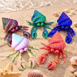 May include: Five articulated crab figurines with spiral shells in various colours including purple, green, blue, pastel pink, and orange. Each crab features multiple jointed legs and two front claws, arranged on a sandy surface near the ocean.