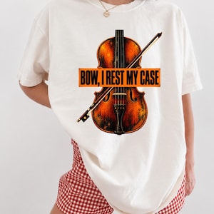Könnte beinhalten: Ein cremefarbenes T-Shirt mit einer Grafik einer braunen Geige und einem Bogen. Über die Mitte verläuft ein orangefarbenes rechteckiges Banner mit dem schwarzen Text "BOW, I REST MY CASE" in fetter Schrift. Die Geige hat eine texturierte Optik.