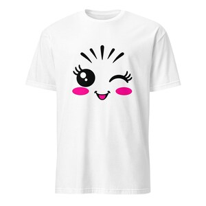 May include: A white short-sleeved t-shirt featuring a centered graphic of a winking face. The design includes black lines for hair, a large black eye with white highlights, a closed winking eye with eyelashes, a small smiling mouth, and bright pink cheeks.