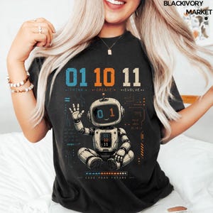 May include: A black t-shirt featuring a graphic of a white robot sitting with one hand raised. Above the robot, the numbers 01, 10, and 11 are printed in blue, orange, and white, with the text THINK, CREATE, and EVOLVE below them.